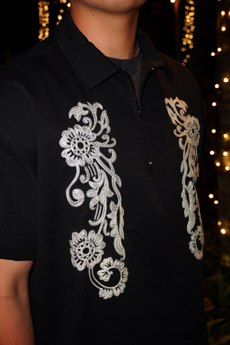 A close-up of a person wearing a black short-sleeve polo shirt with intricate white floral embroidery on the front, set against a dark background with warm string lights.