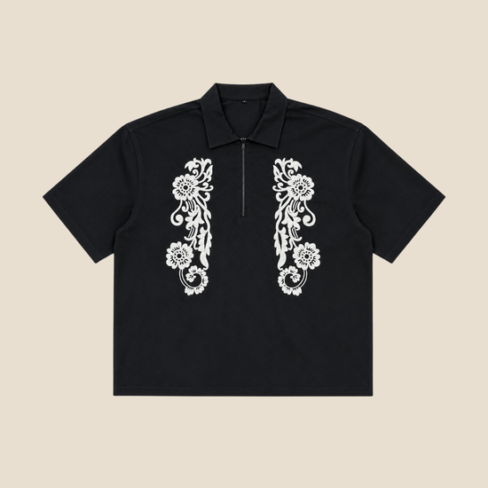 A black short-sleeve polo shirt with a half-zip collar and white floral embroidery on the chest, displayed flat against a solid beige background.