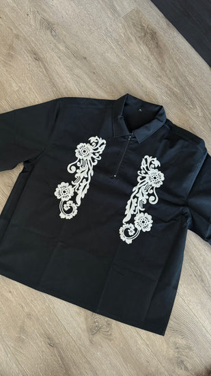 A black short-sleeve collared shirt with white floral embroidery on the chest, laid flat on a light brown wood floor.