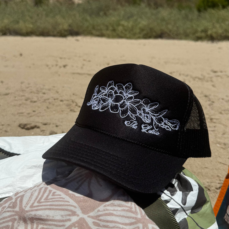 Foam trucker hat with embroidered Plumeria flower laid on top of beach bag at the beach.