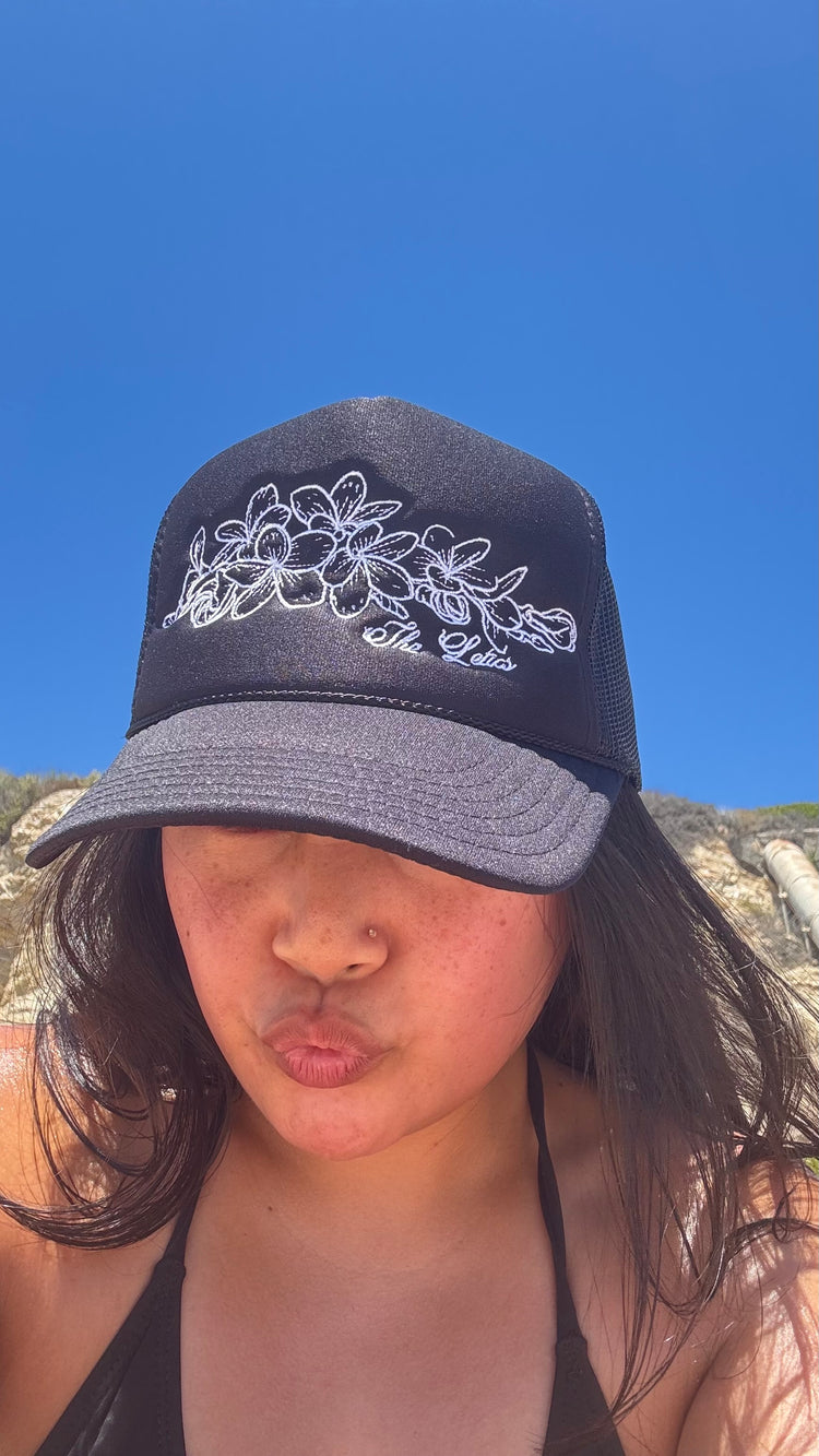 Person wearing a black foam trucker hat with white floral embroidery against a blue sky at the beach.